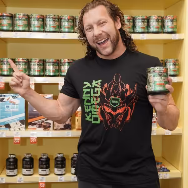 Kenny Omega smiling and holding a container of Total War pre-workout in a supplement store