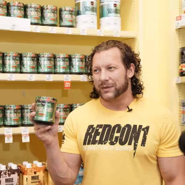 Kenny Omega holding a container of Redcon1 Total War pre-workout in front of a supplement display
