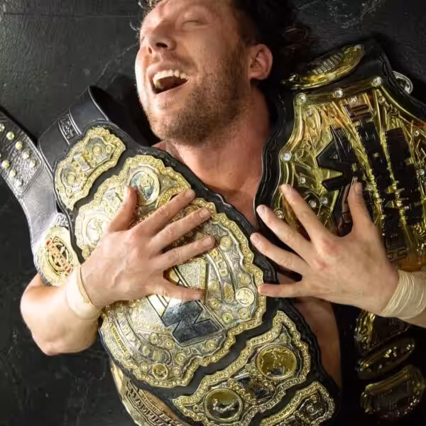 Kenny Omega lying on his back, laughing while draped in multiple gold championship belts